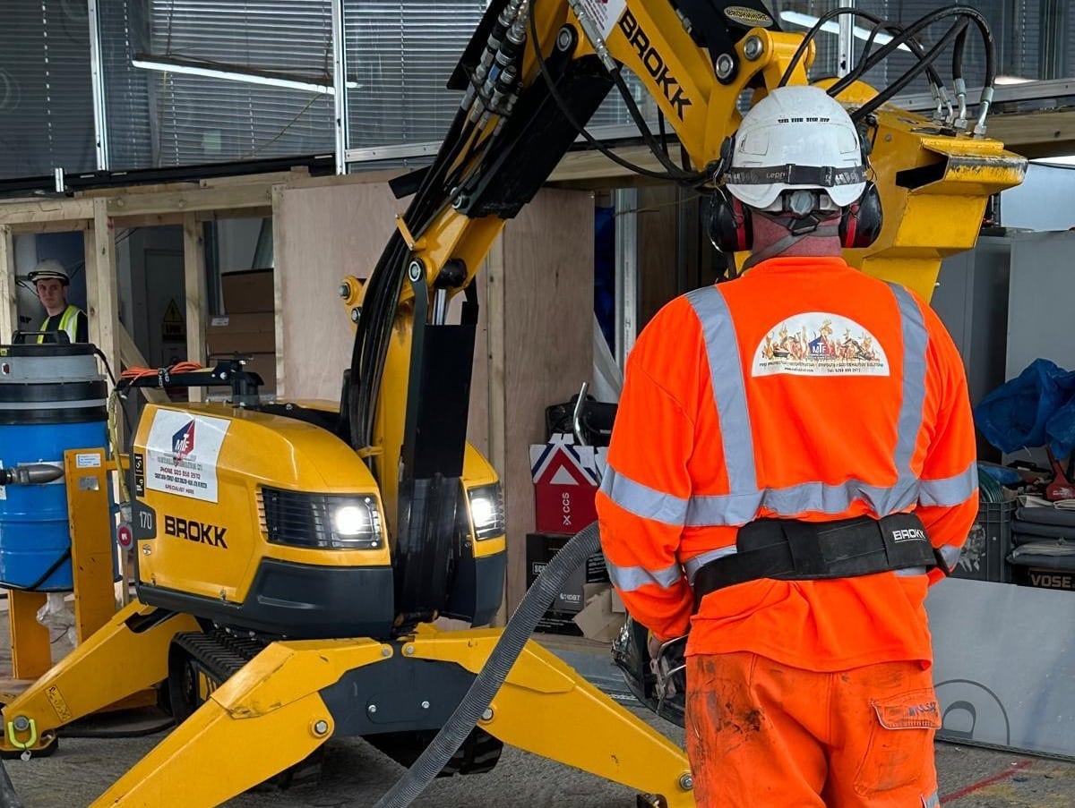 A construction worker in orange gear operates a yellow demolition machine indoors.