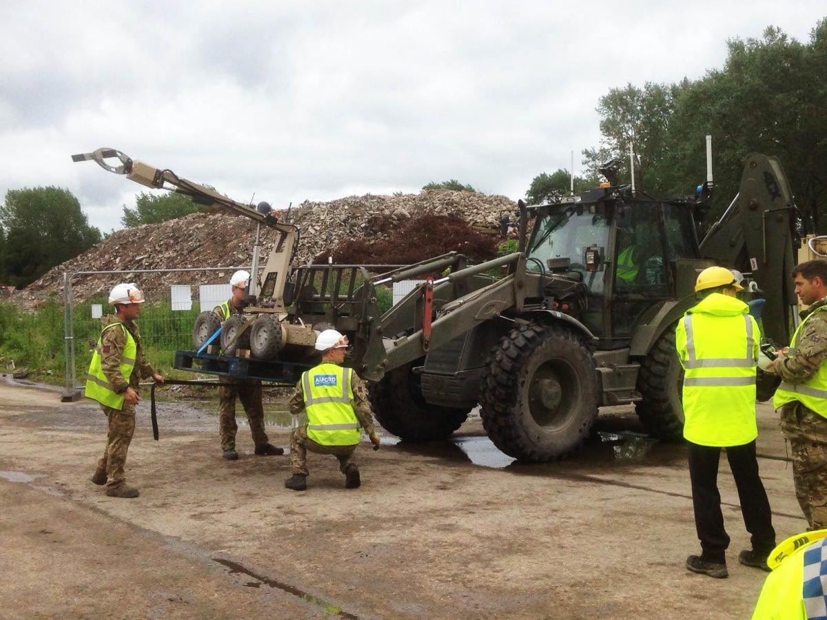 Workers in safety gear operate machinery at a construction or demolition site with rubble in the background.