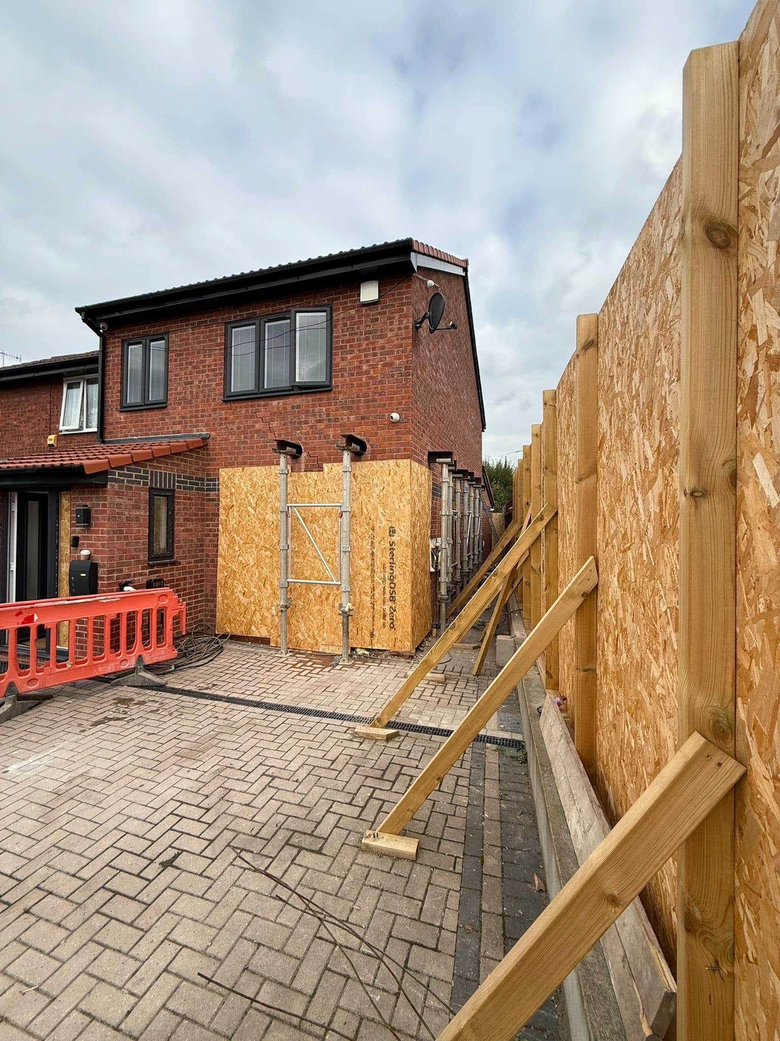 Wooden temporary supports and barriers beside a brick house under construction on a cloudy day.