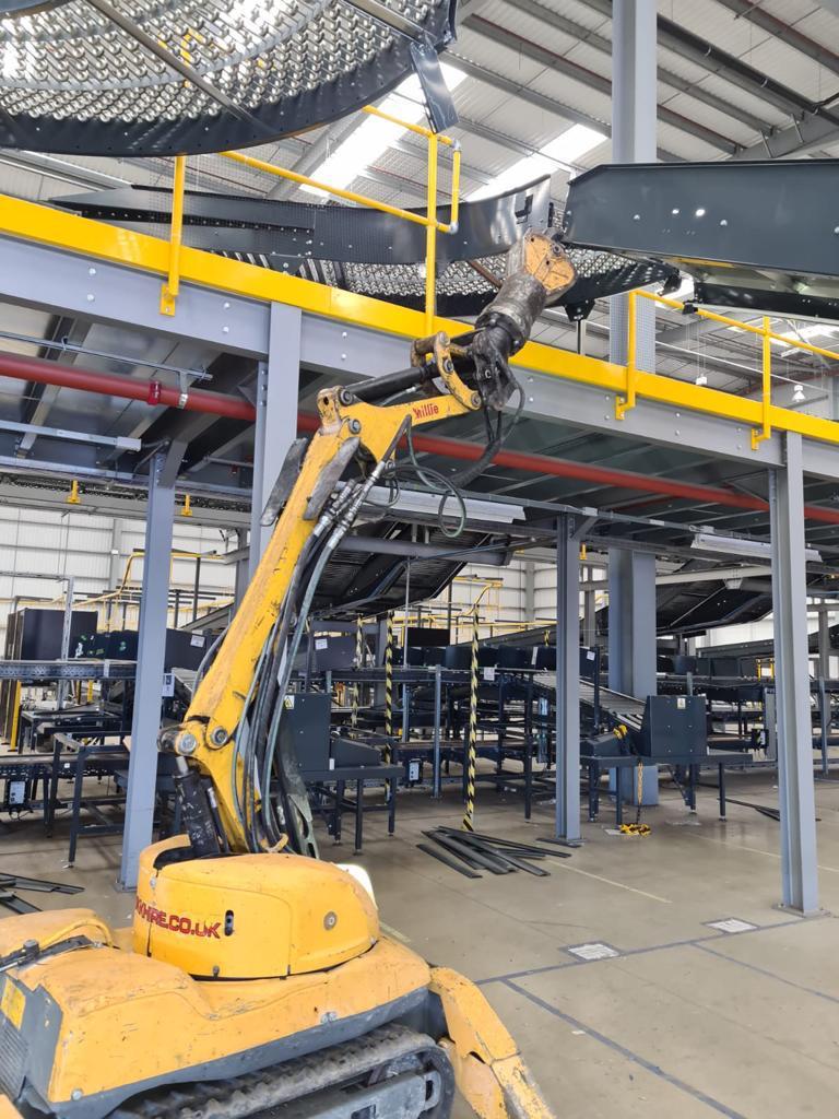 A yellow demolition robot operates inside an industrial warehouse with metal structures.