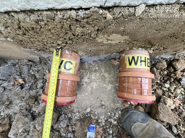 Two labeled drainage pipes, "WC" and "WHB," in soil with a measuring tape beside them.