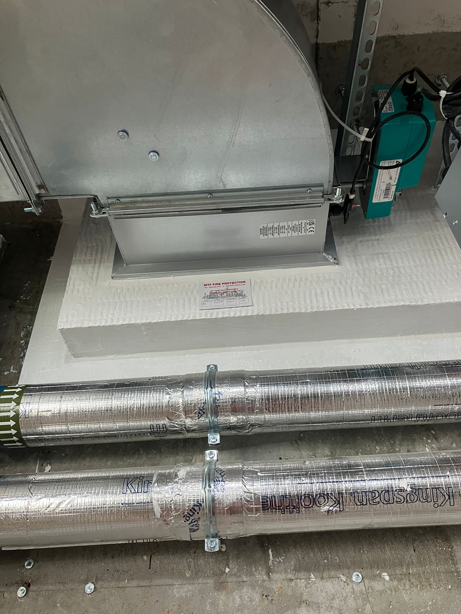 Metal pipes and ductwork on a white firestopping barrier in a mechanical room.