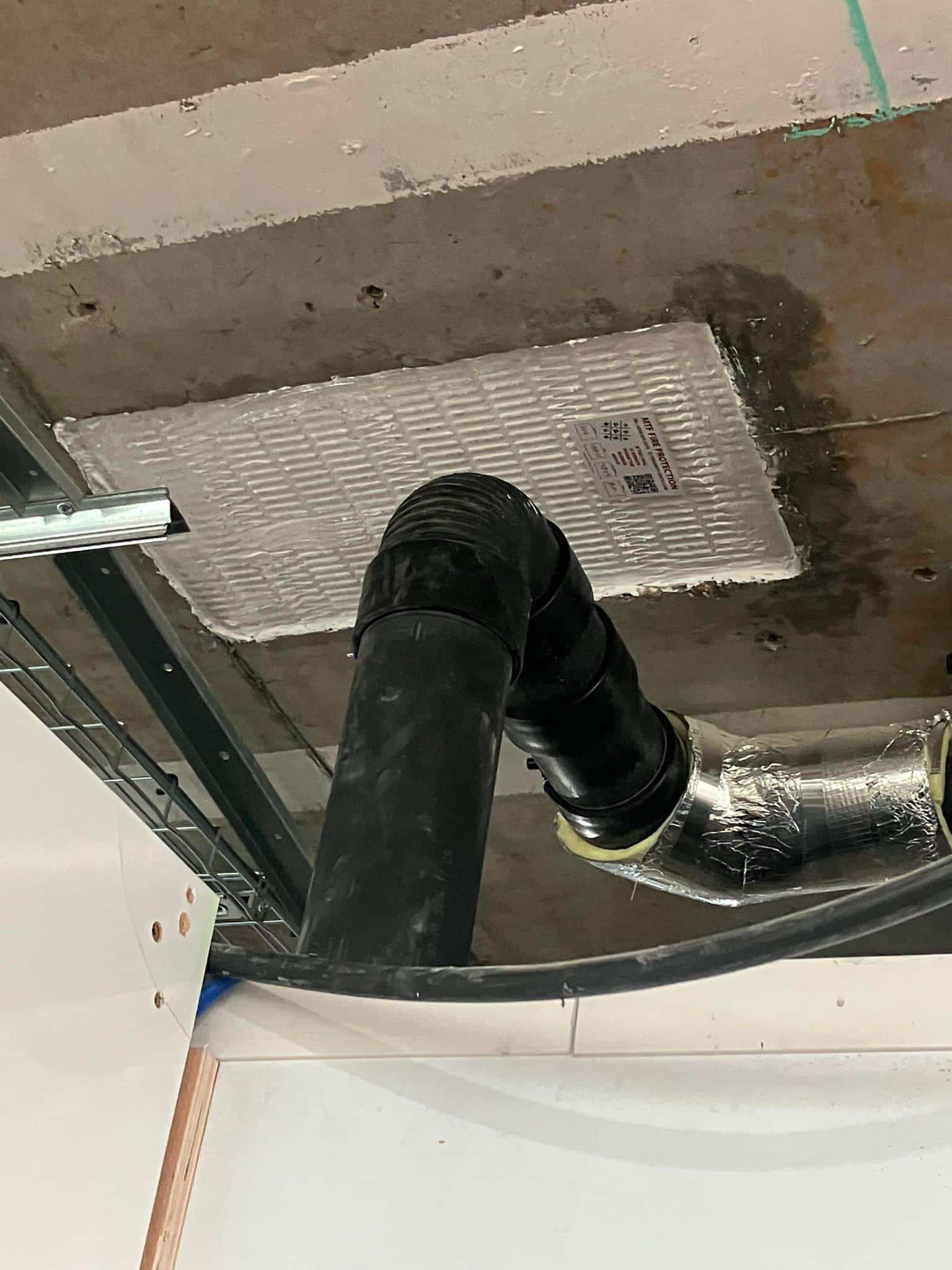 Black pipes and a duct run below a firestopping board installed in a concrete ceiling.
