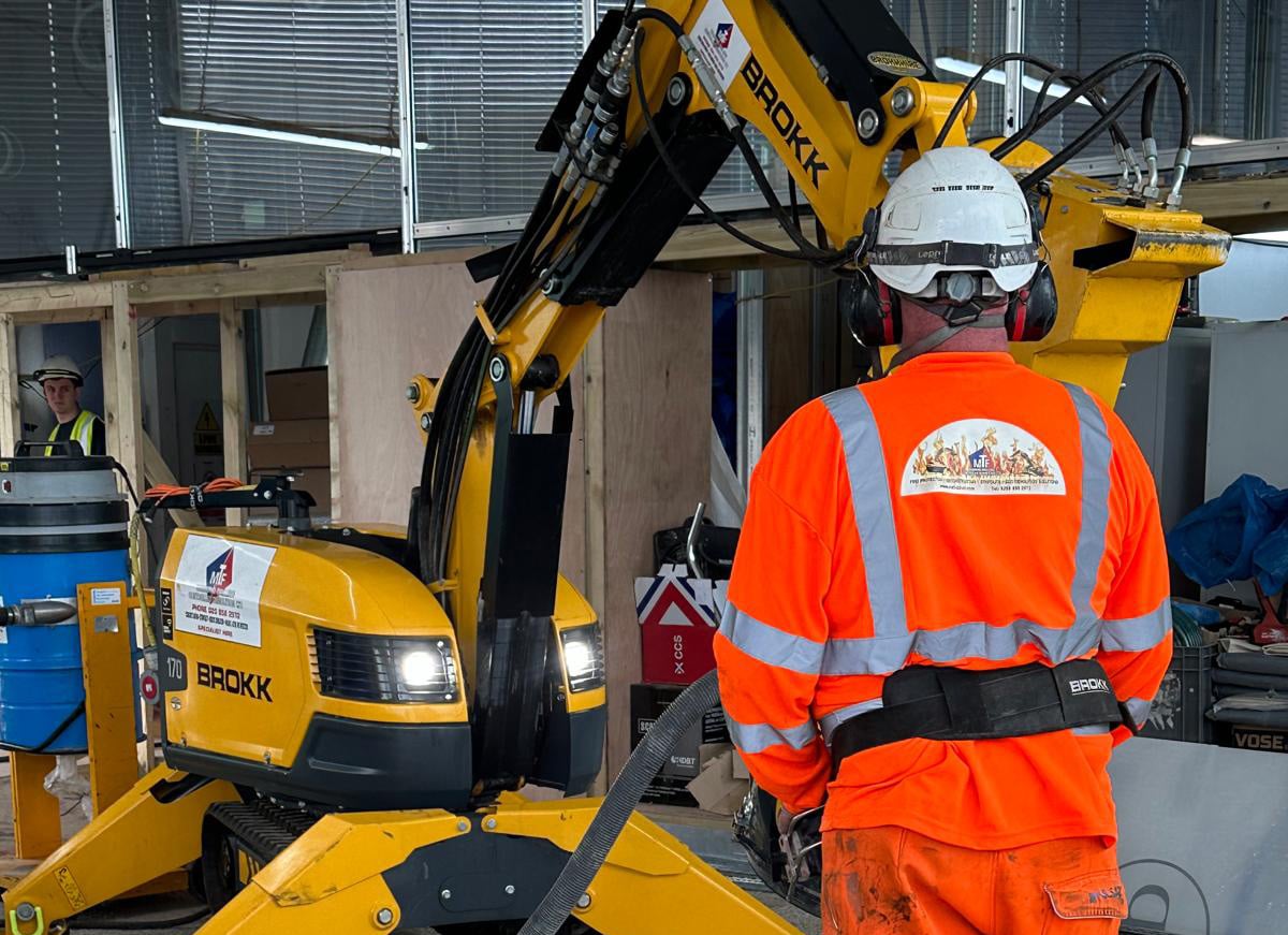 Worker in orange safety gear operating a yellow construction machine indoors.
