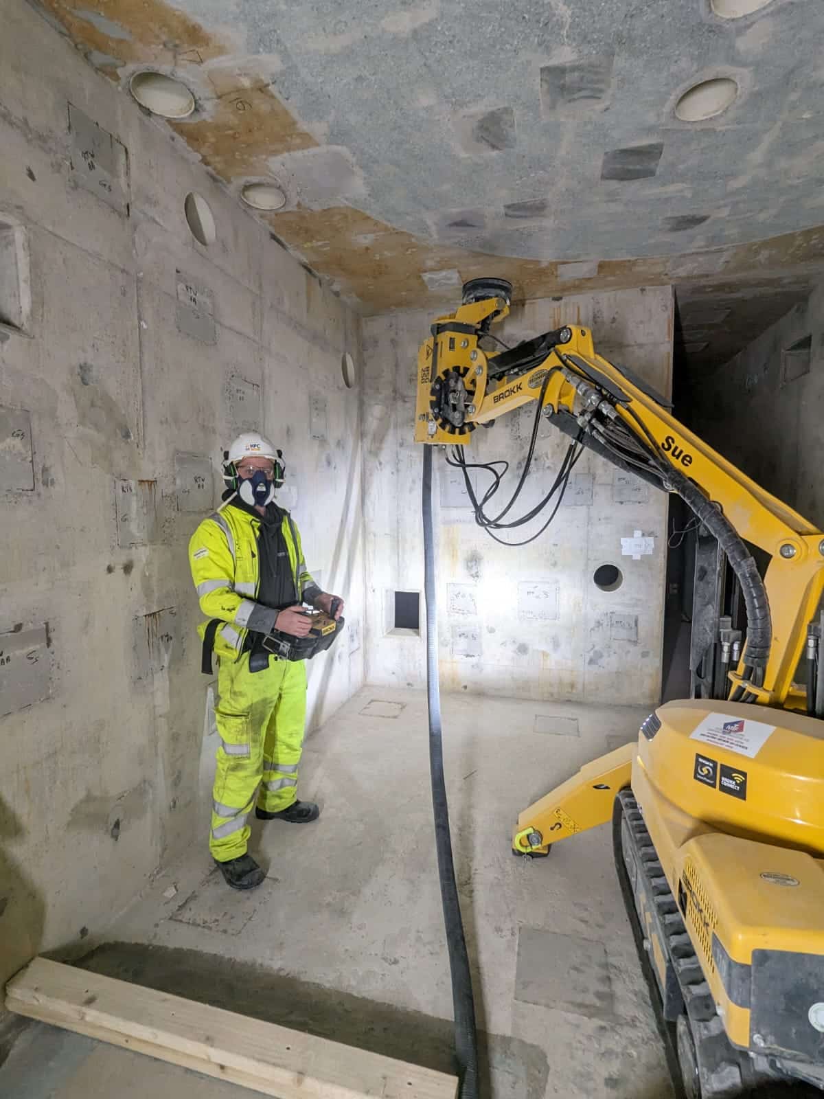 Worker in safety gear operates a yellow construction machine inside a concrete room.