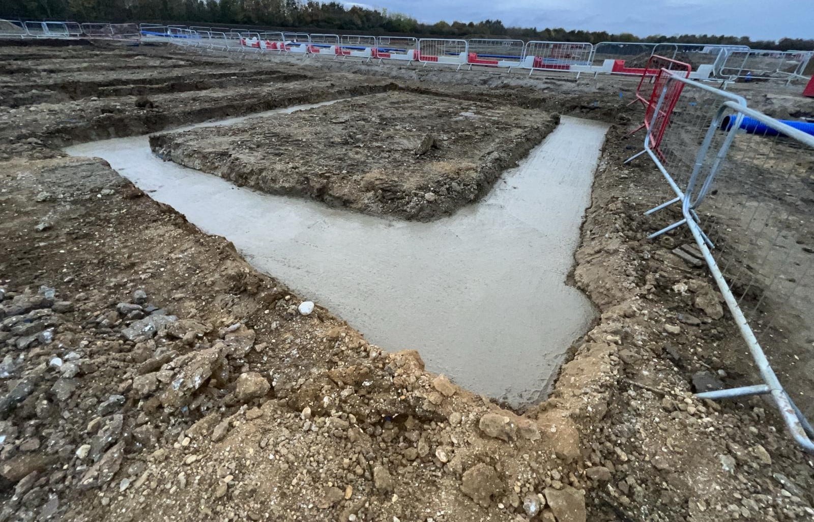 Concrete footing poured in a trench at a construction site with soil and safety barriers nearby.