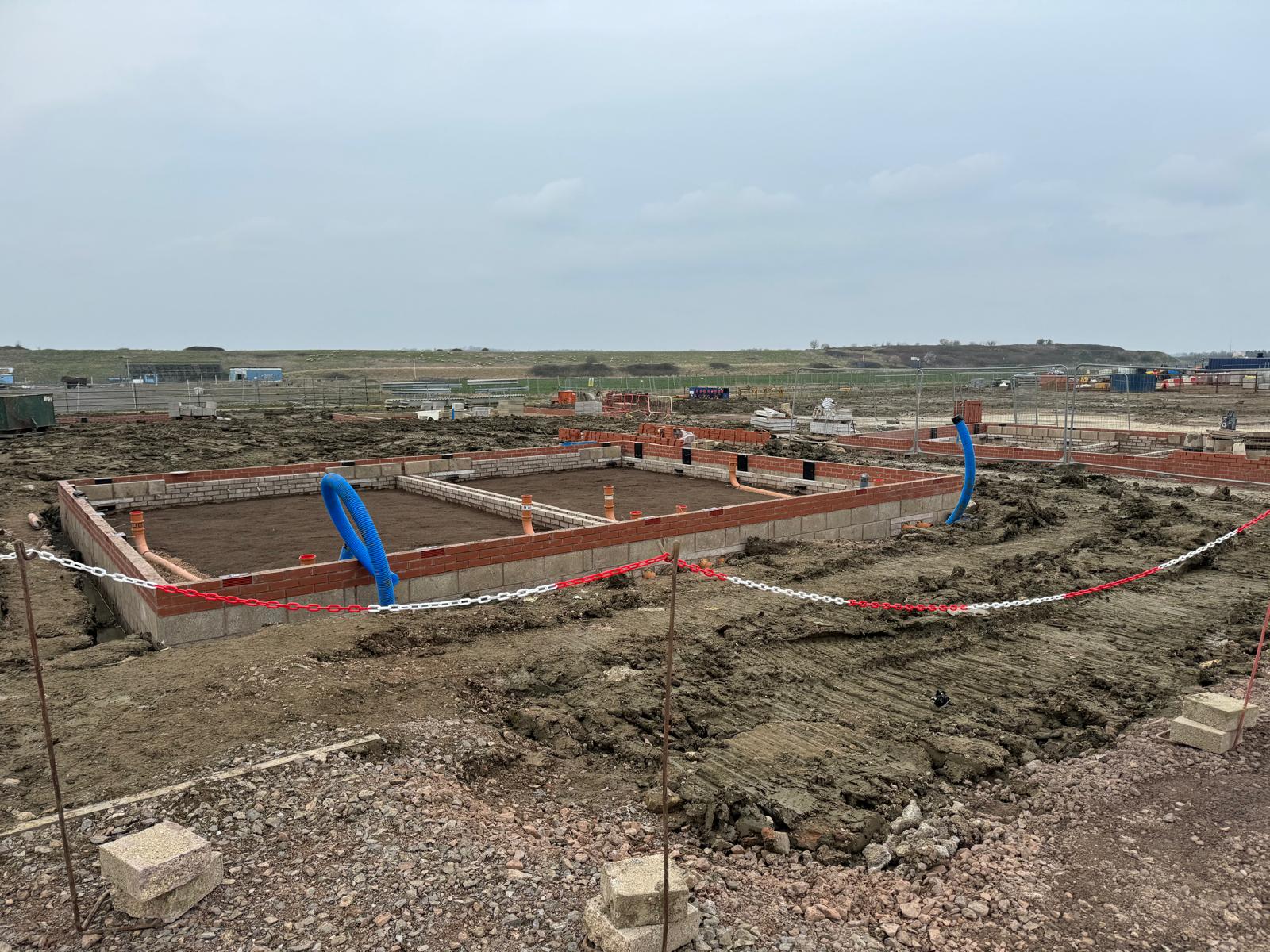Building foundations under construction on a muddy site with pipes and red bricks visible.