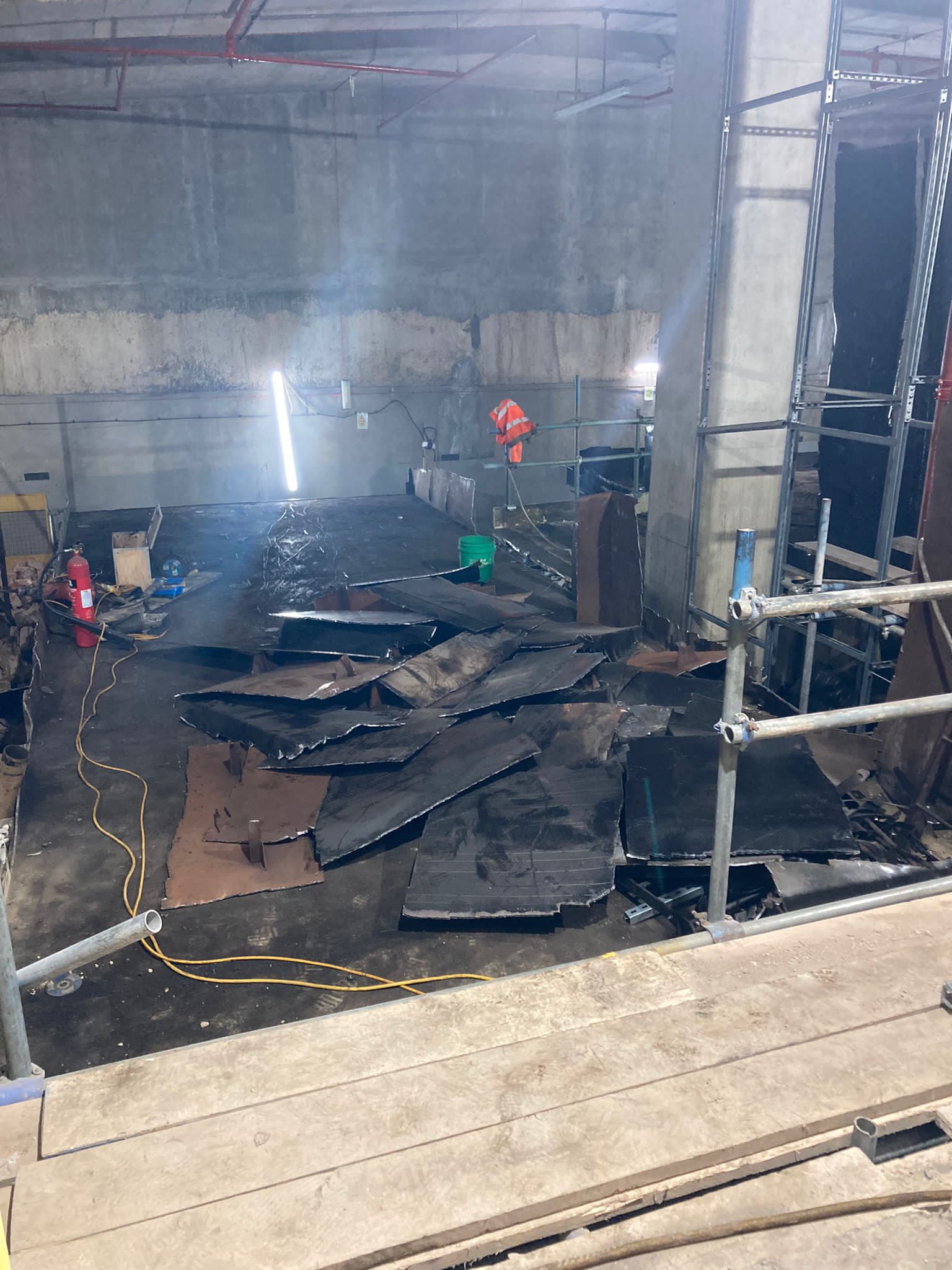 Construction site with debris and metal sheets scattered on the floor, scaffolding visible on the right.