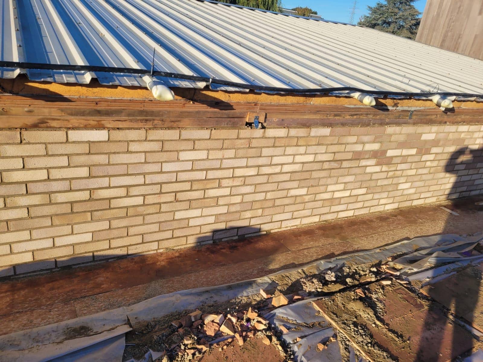Brick wall under a corrugated metal roof, with construction materials on the ground.