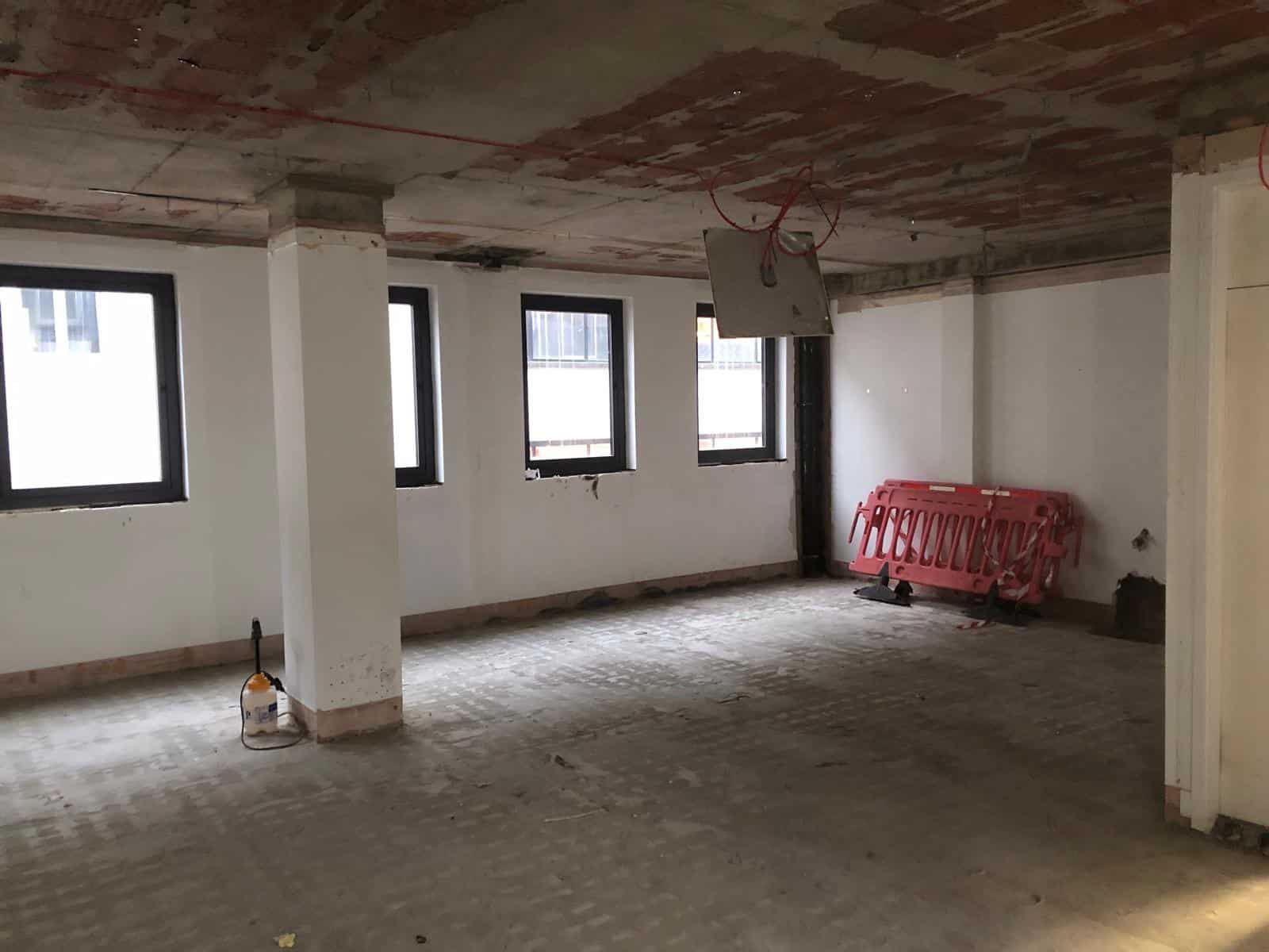Empty room under renovation with three windows, exposed ceiling, and construction materials in the corner.