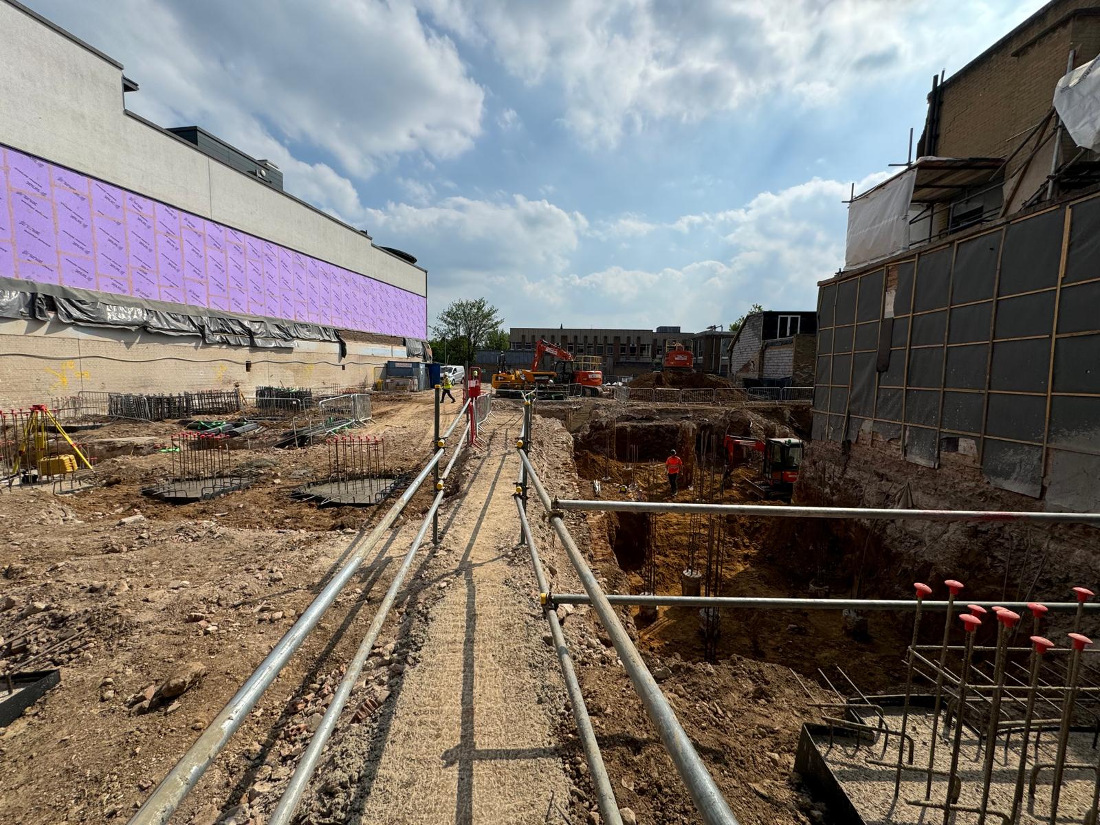 A construction site with dirt, steel bars, and a fenced walkway under a partly cloudy sky.