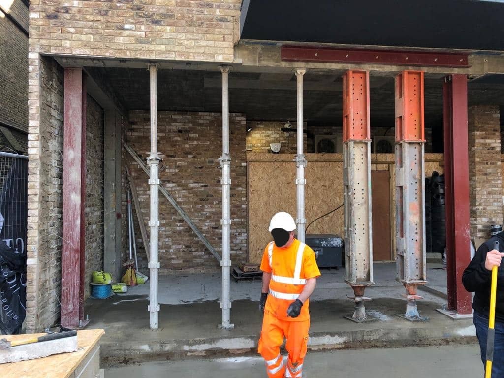Construction worker in orange safety gear stands in front of steel support beams at a building site.