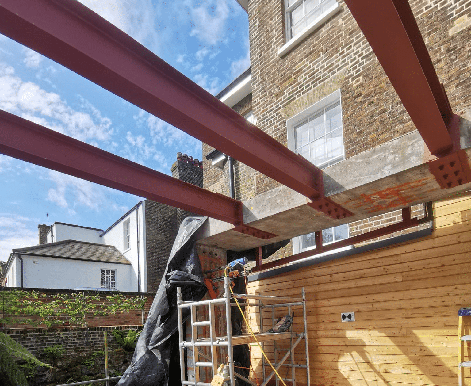 Red steel beams installed on the exterior of a brick house, with scaffolding below.