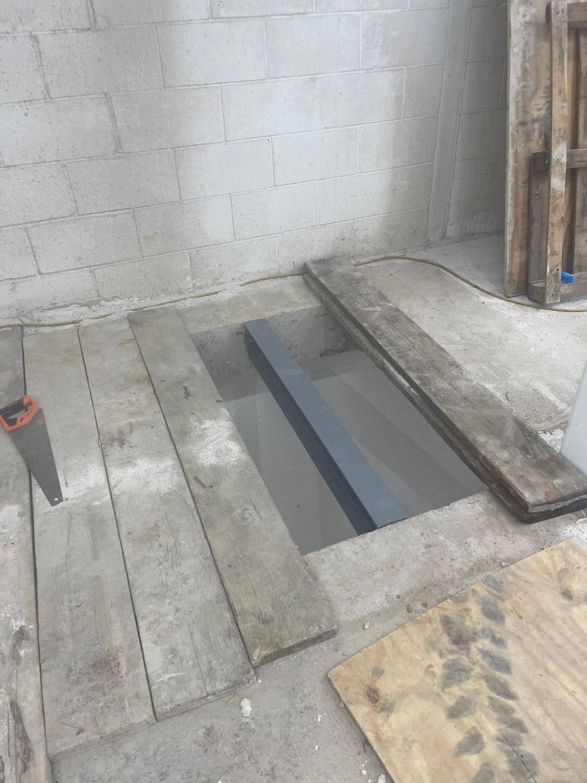 Wooden planks covering a rectangular hole in a concrete floor by a cinder block wall.