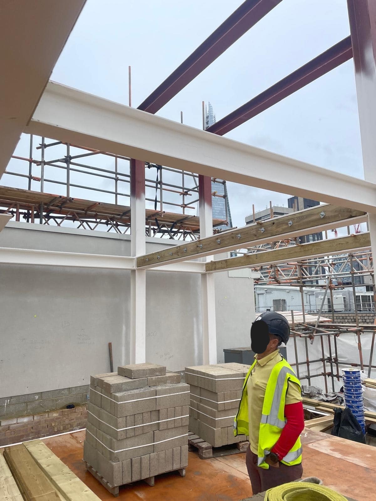 Construction worker in safety gear stands near steel beams and stacked bricks at a building site.